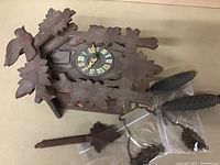 Front view of antique wooden cuckoo clock showing carved bird, leaves, dial with Roman numerals, and clock hands on a chalet-shaped housing.