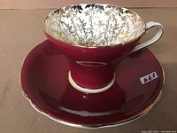 Side view of the cup sitting on saucer showing burgundy color and gold leaf pattern inside cup.