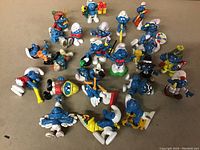 Photo showing a varied collection of vintage Smurf figurines on a tan surface. The figurines depict several Smurfs in different poses and outfits, including musical instruments, gifts, and various accessories.