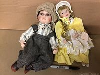 Two porcelain dolls on cardboard background, boy doll wearing beige knit cap, striped shirt, dark overalls; girl doll in yellow dress and white bonnet