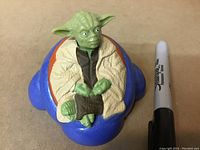 Top view showing detailed Yoda figure in green with beige cloak seated on blue Magic 8-Ball base next to a Sharpie for scale.