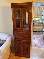 Front view of tall wooden cabinet showing glass upper doors and front drop-front compartment with lower double doors