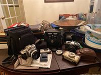 Full lot of cameras, binoculars, bags, and accessories shown on table.