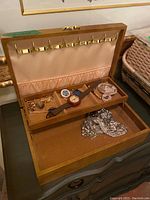 View of open wooden jewellery box with soft beige interior containing assorted costume jewellery, mechanical watch, brooches and silver-tone accessory.