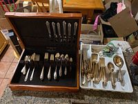 Silver plated flatware arranged in wooden storage box and additional plated cutlery in white plastic kitchen drawer insert on table.