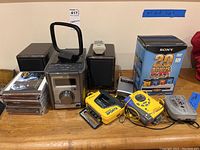 Wide view of all electronics including JVC stereo system, speakers, Sony Walkmans, portable radio, stack of CDs, and box of Sony blank DVDs.