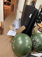 View of two black exercise mats standing vertically with carrying handles, one camouflage green exercise ball, pedaler in packaging with instructions on a box
