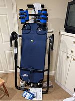 Front view of folded inversion table showing blue backrest, ankle restraint pads and black frame