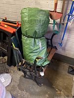 Cast iron bench with wooden slats visible, rolled green tarp strapped and placed on bench seat, surrounded by garage items, taken to show main furniture items in lot.