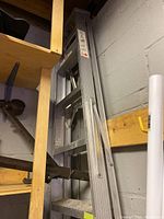Folded 8-foot aluminum stepladder stored against wall next to wooden shelving and a concrete block wall.
