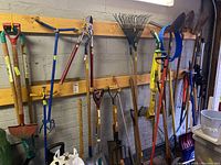 Wide view of multiple long-handled shovels, rakes, forks and edging tools hung on wall rack