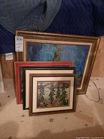 Four framed paintings and prints stacked and leaned against a wall, showing various sizes and frame styles.