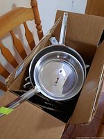 Photo of stainless steel saucepan with lid inside cardboard box on chair.
