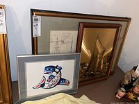 Three framed artworks displayed, including a colorful indigenous style print, a maritime sailing ship print, and a metal etching with ship rigging details.