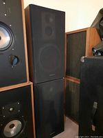 Two tall, wood frame Technics speakers with black front fabric covers stacked vertically in a room corner with other audio speakers visible nearby.