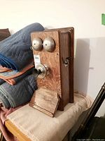 Side view of the vintage wooden hand cranked box telephone showing the metal bells, crank, and mouthpiece with natural wood grain finish