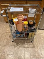 Front angled view of the three-tier Wiretech wire kitchen cart showing metal wire shelves, attached handle, and wheels. Contains various kitchen items that are not included in the lot.