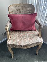 Pair of cane arm chairs set near window, showing carved wood frame, woven cane back and sides, apricot-beige patterned seat cushion, and rectangular muted reddish-pink back cushion.