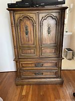 Front view of wooden armoire showing ornate double doors with decorative metal handles above and two large drawers below with matching handles.
