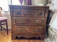 Photo showing a 3 drawer wooden bureau with detailed wood carving, brass colored decorative handles, and a scalloped base.