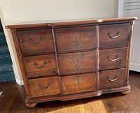 Photo shows a wooden bureau with three drawers. Each drawer features metal handles and painted floral designs. The piece has a curved front and rounded feet, with a medium brown finish.