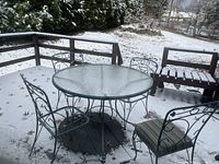 Outdoor dining set on a snowy deck including a round black metal table with glass top and four black metal chairs with removable cushions