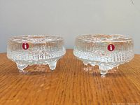 Pair of two Iittala Ultima Thule crystal candleholders with original red Iittala stickers facing front, displayed on wood surface.