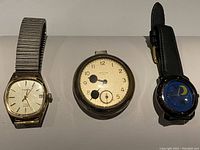 Three watches displayed side by side: silver pocket watch, gold-tone Cardinal wristwatch, and black leather strap wristwatch with moon/star design.
