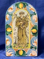 Front view of ceramic wall plaque depicting St Francis in robe with bird and lamb, surrounded by colorful ornamental border