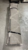 Rolled Thomasville Thatcher gray rug secured with black straps showing backing and fringed edge.
