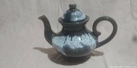 Side view of a ceramic teapot showing its blue and white glaze with rounded body, curved spout, and attached handle.