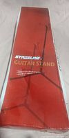 Box packaging for Stageline Guitar Stand showing the product name, universal fit for all guitar styles, folding neck support, and safety strap features.