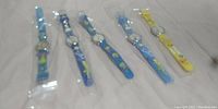 Five children's wristwatches individually packaged in plastic sleeves, laid out side by side. Straps are colorful and feature cartoon characters.