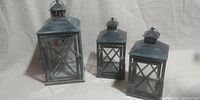 Front view of three metal lanterns of different sizes arranged side by side, showing aged finish and glass panels.