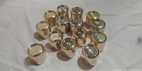 14 small polished brass cylindrical containers, arranged in a triangular shape with lids on, on white surface.