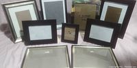 Nine assorted picture frames arranged on a surface showing variety in sizes and colors including silver, black, and gold tones. All have glass fronts and backing boards. Some frames have visible wear and tear.
