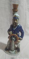 Front view of vintage ceramic sailor decanter, showing sailor figure steering ship's wheel with cork stopper on top.