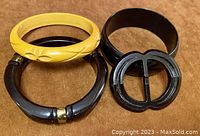 Three Bakelite bangles and one black Bakelite ribbon/belt buckle, shown flat on brown surface.