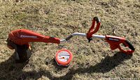 Full view of the Black & Decker GH700 GrassHog 14" trimmer/edger with orange body, silver metal shaft, and black trimming head. Unopened roll of trimmer line placed beside it on grass.