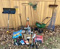 Full view of garden tools and supplies arranged on ground against wood fence, showing shovels, rakes, hand tools, gloves, soil bags, tomato cages, watering can and seeder.
