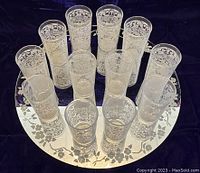 Top-down view of set of 12 tall glasses arranged standing on decorative round mirror with floral border.