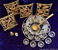 Full set arranged: punchbowl with cups in wire holder, ladle, four decorative gold leaf season plaques, and cut glass salt and pepper shakers with gold tops on dark fabric background.