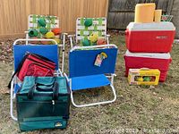 Overview photo showing two blue Flamingo low beach chairs, two aluminum lawn chairs with woven seats, Bocce ball set box, coolers and patio string lights on grass