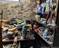 Workspace with shelves stacked with various small tools, containers, and hardware supplies along with cluttered paperwork and cloths on a wooden desk beneath a stone window wall.