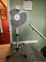 Cypress electric pedestal fan positioned near stairway and wall, showing entire fan with adjustable pole and three-legged base.