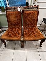 Photo showing front view of two large handcrafted side chairs with burnt orange velvet patterned fabric seats and backs. Ornate carved wood frames with curved legs and decorative trim at top.