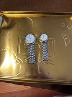 Photo showing two wristwatches, one larger men’s model and one smaller ladies’ model, both with silver metal link bracelets and white faces with Roman numeral markings, placed on a metallic surface.