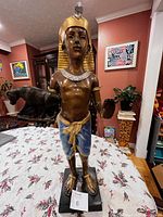 Full frontal view of an Egyptian style pharaoh statue holding a black panther figurine standing on a rectangular black base, on a table with floral tablecloth, in a living room with art on walls.