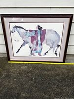 Front view of the framed watercolor painting leaning against exterior wall showing woman in traditional attire with horse, with measuring tape showing approximate size.