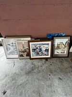 Photo showing six framed paintings and pictures arranged leaning against a wall on a concrete floor.